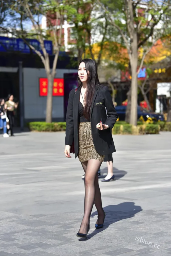 都市时尚街拍:一位身穿黑色西装、金色亮片短裙、搭配黑色丝袜和高跟鞋的女性,在宽阔的城市广场上自信行走,展现优雅与时尚。